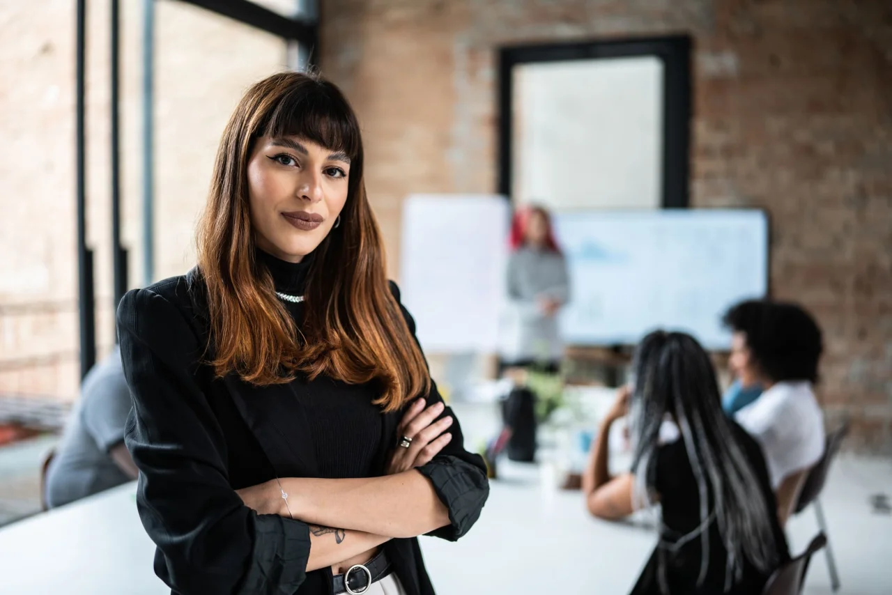 Confident female business leader standing with arms crossed in modern meeting room with team in the background.