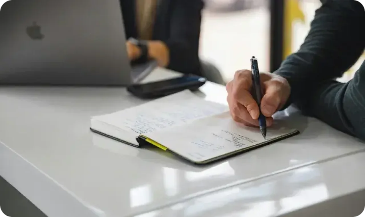 A man writing notes in a notebook and woman working on PC