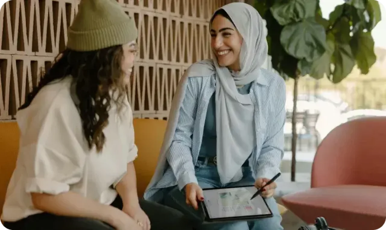 Two women sitting and smiling while discussing something on a tablet; one wears a hijab, the other a beanie