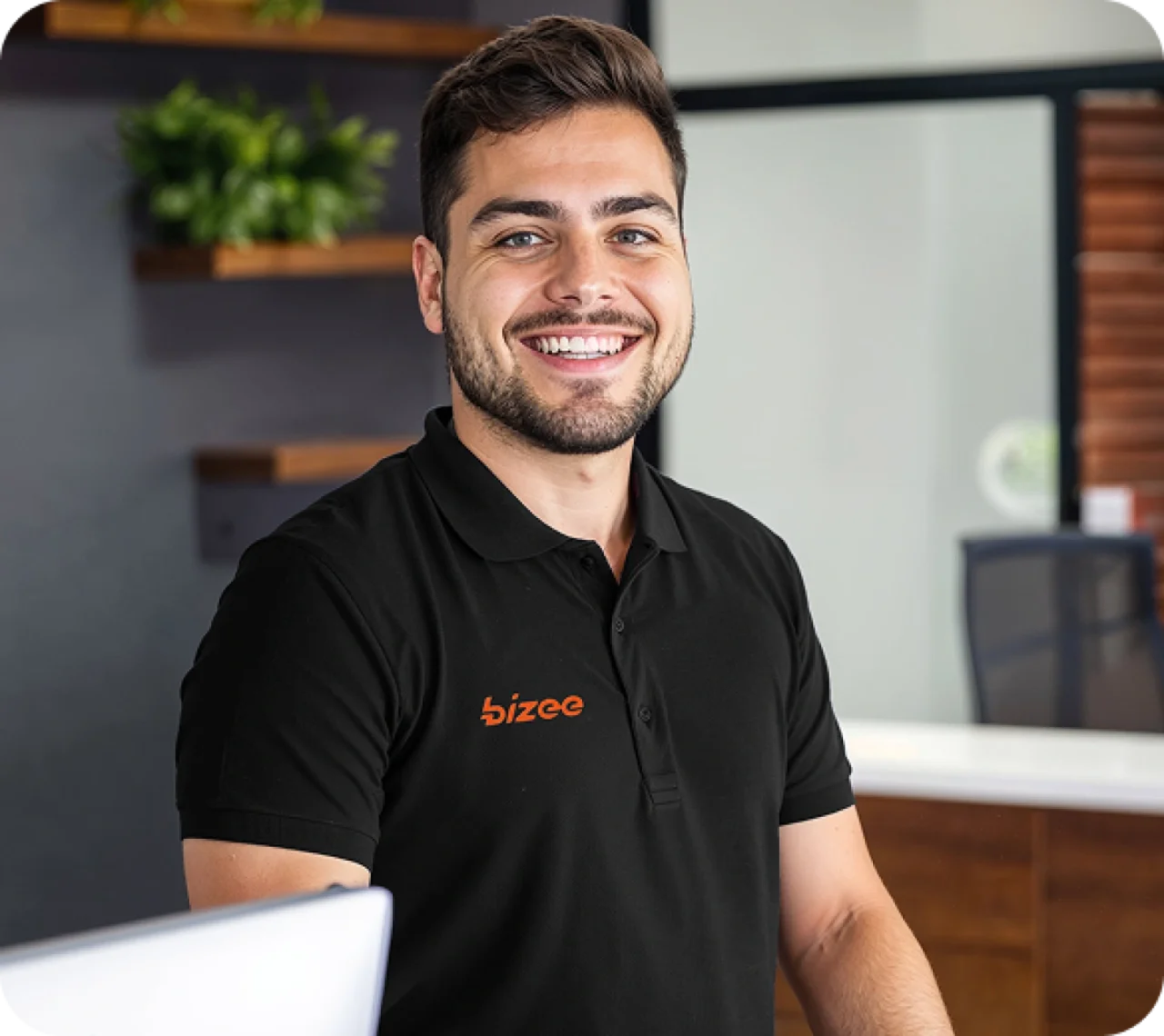 Smiling male customer support representative wearing a black Bizee polo shirt, seated at a modern office reception desk with wooden accents and green plants in the background.