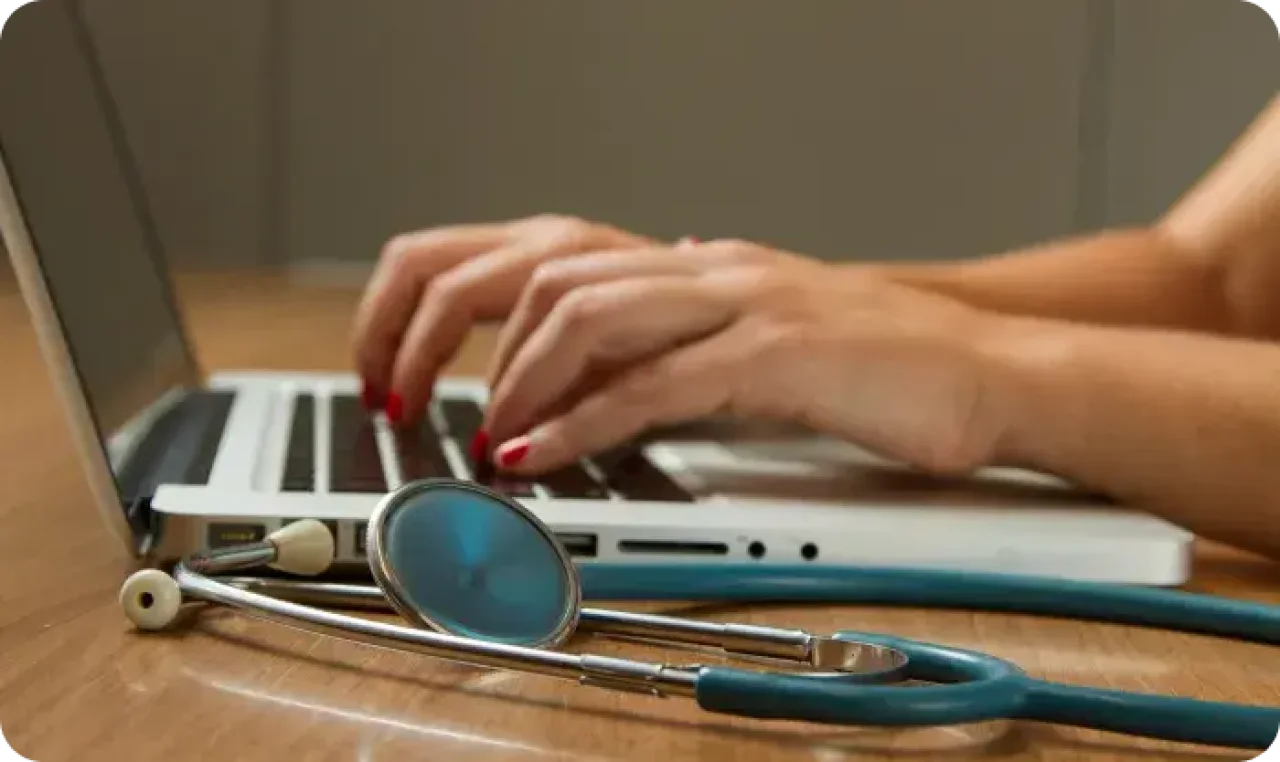 close-up-of-hands-typing-on-a-laptop-with-a-stethoscope-resting-on-a-wooden-desk-indicating-a-medical-or-healthcare-setting