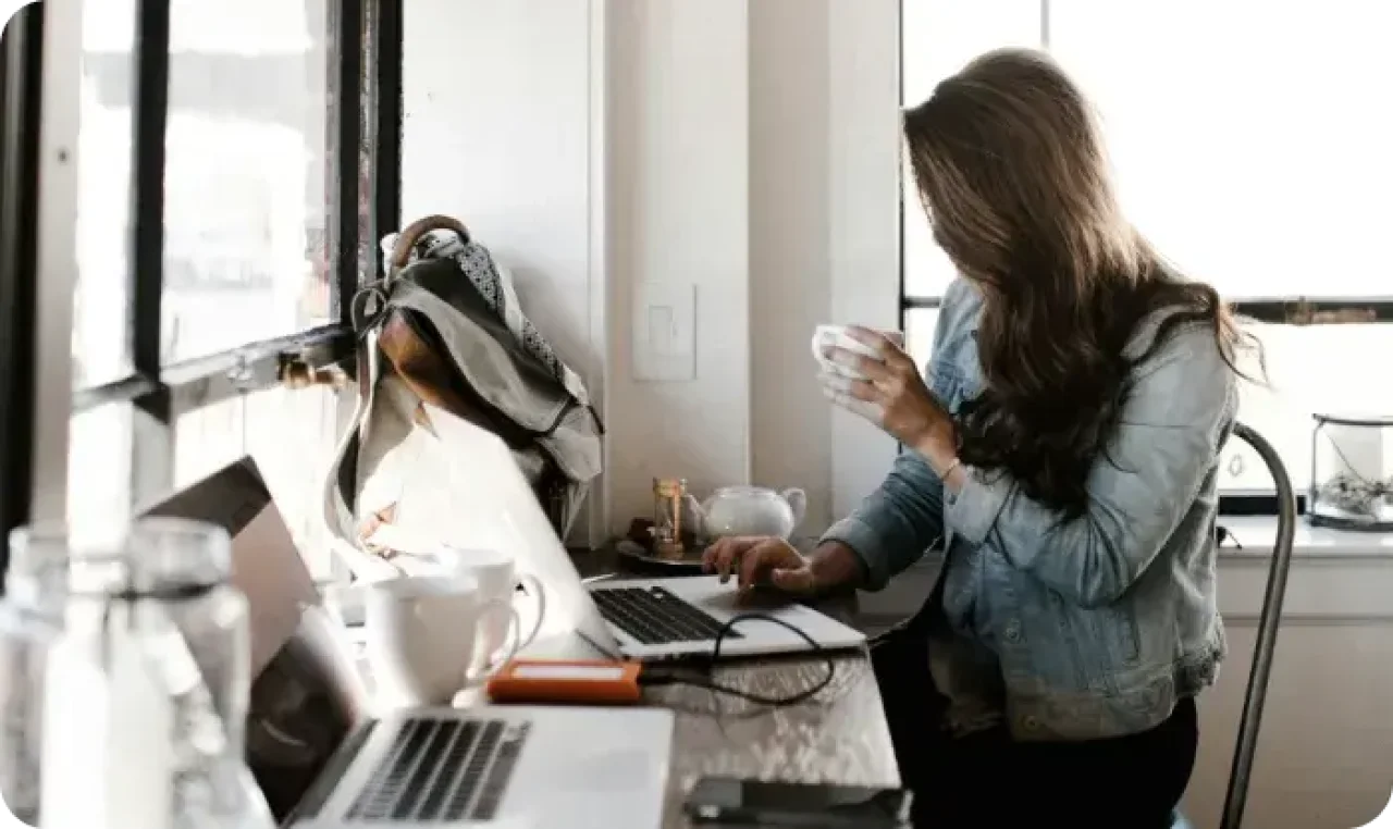 A woman seated at a desk, focused on her laptop, with a cup of coffee beside her, creating a productive workspace.