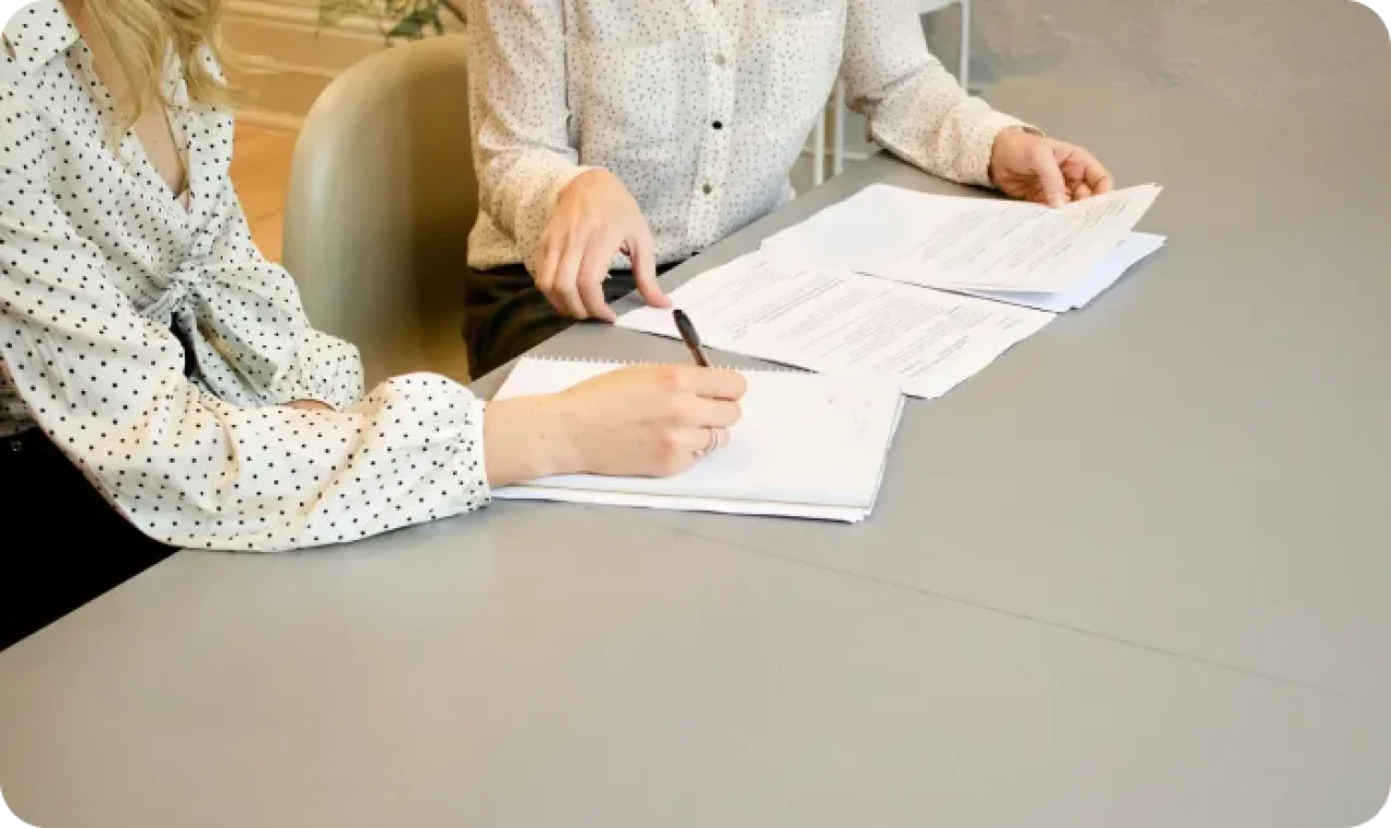 Woman signing on white printer paper beside woman about to touch the documents