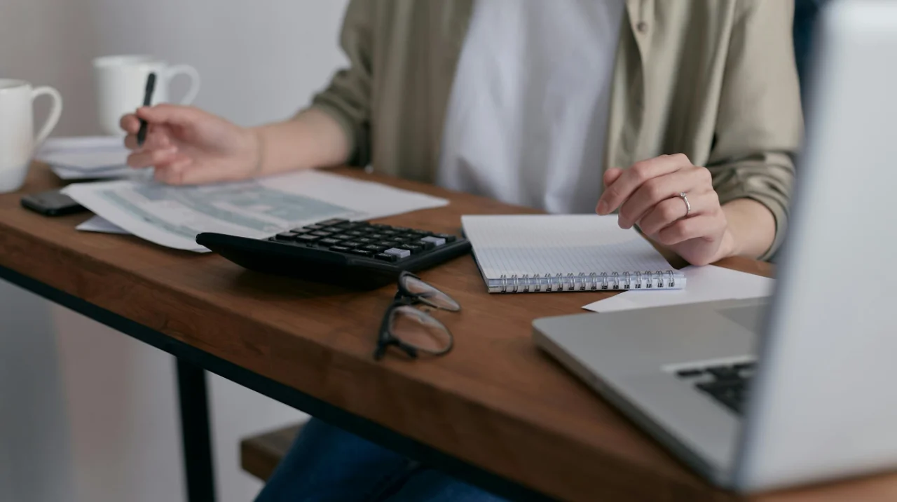 Person working on tax documents with a calculator, notebook, and laptop at a desk, preparing financial records
